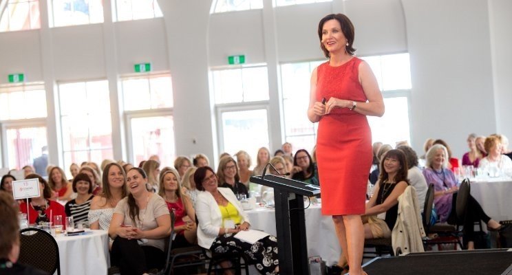 Woman with long light brown hair pinching the bridge of her nose with eyes closed, wearing a light pink top, against a light gray background